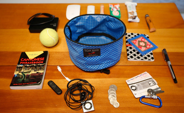 A Large Travel Tray surrounded by various accoutrements: a Cantonese Phrasebook, a USB keydrive, a charging battery, a dongle, a pair of earbuds and MP3 player, a bit of change, a carabiner and key, a Metrocard, a pen, a notepad, a tube of lotion, some snacks, and a tennis ball.
